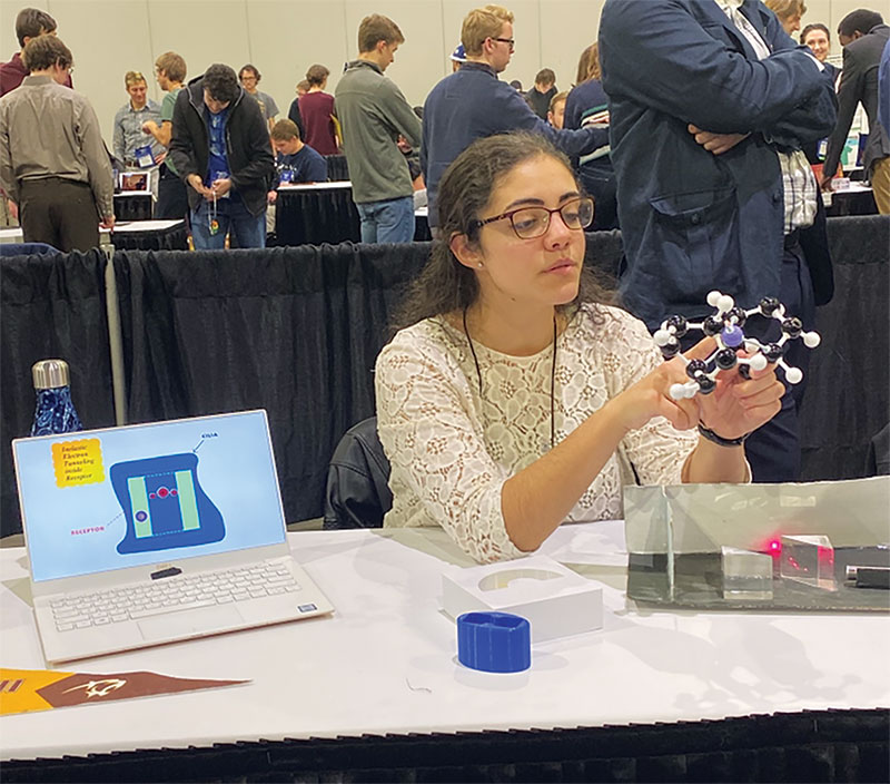 Giuliano presents her work on smell at the 2019 Physics Congress. She is holding a model of a molecule that can represent either nickelocene or ferrocene—molecules with similar structures but different smells. The laptop and laser demo illustrate inelastic electron tunneling, and the white and blue pieces on the table are 3D-printed olfactory receptors.