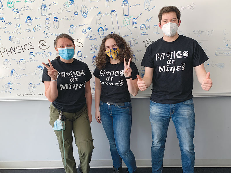 Mines SPS officers (L-R) Olivia Jackson, Elizabeth Buchheim, and Connor Hewson sport newly designed shirts in front of the “Physics at Mines” ghost sketch in the lounge.